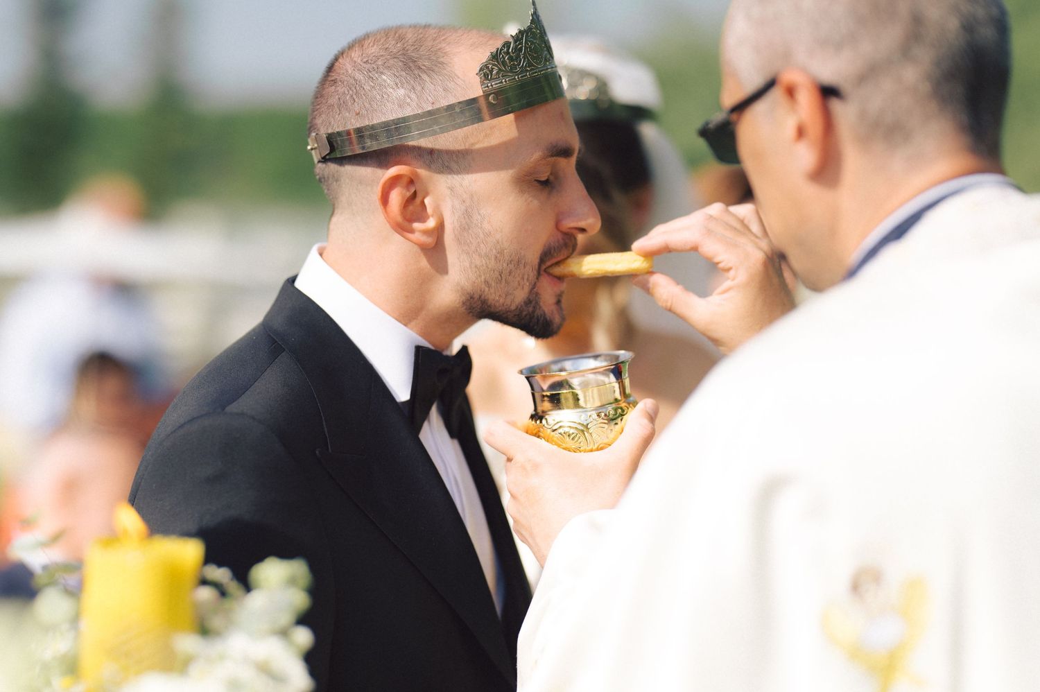 Two people in formal attire share cake at an outdoor wedding celebration.