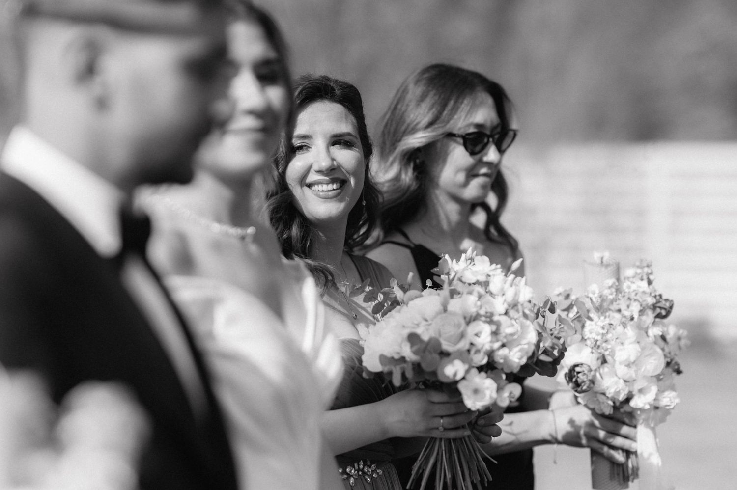 Black and white image of a bridal party sharing a joyful moment during a wedding ceremony, with elegant floral bouquets.