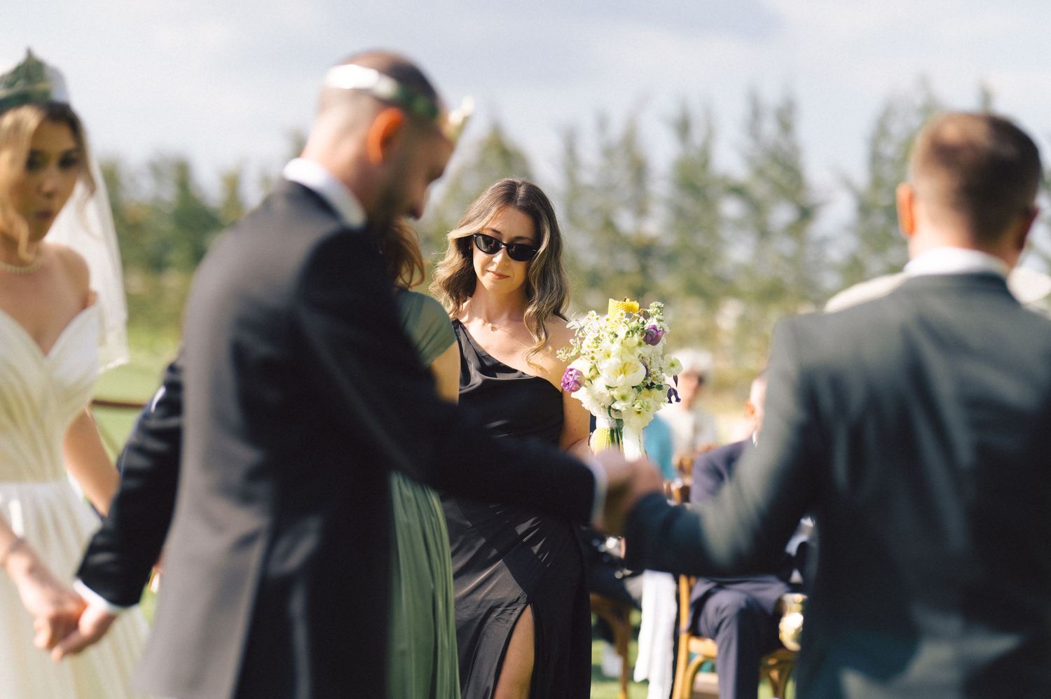 People gathered at an outdoor wedding ceremony with guests wearing formal attire in a garden setting.