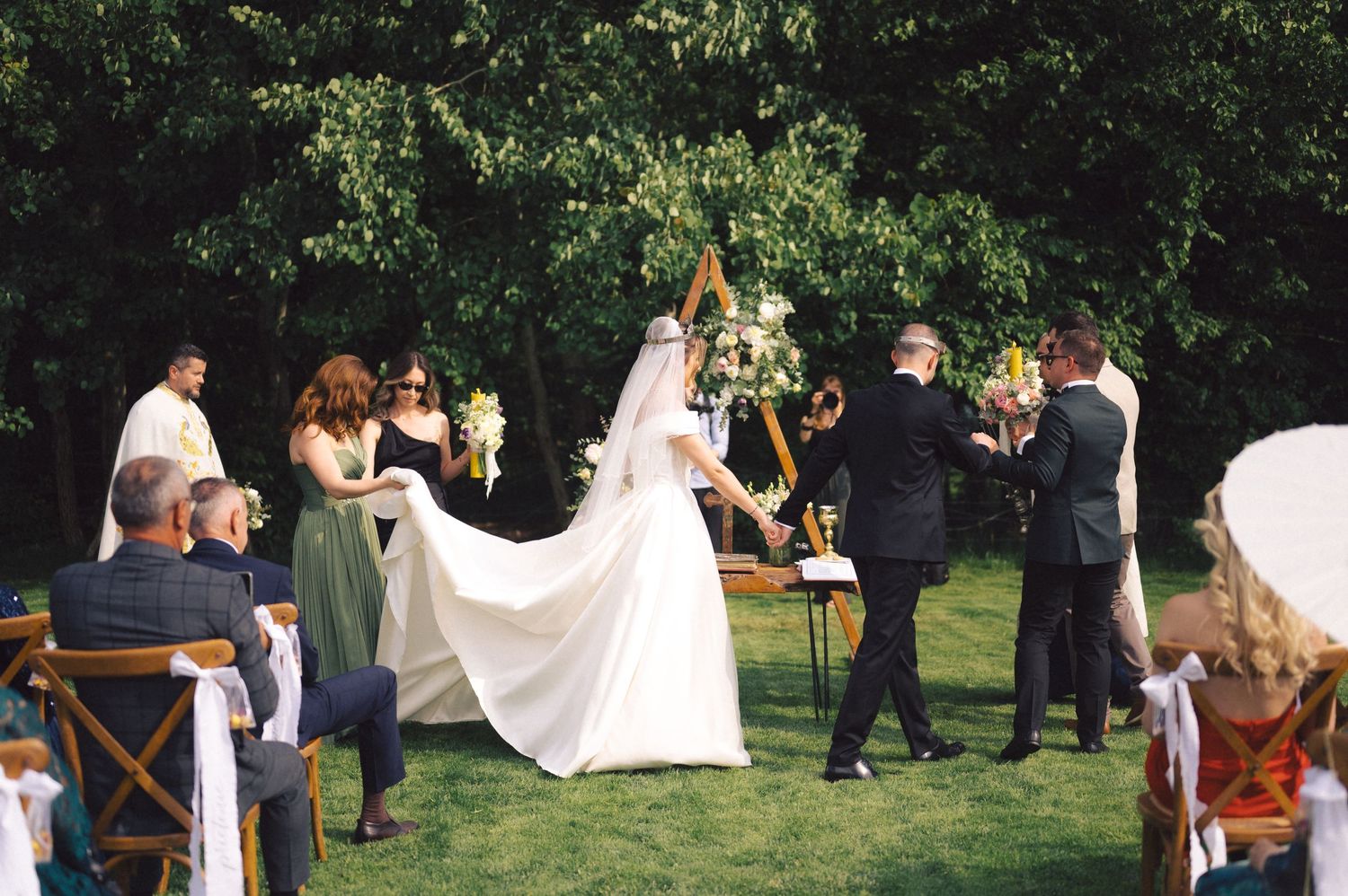 A beautiful outdoor garden wedding ceremony with attendees seated on wooden chairs as flowing white dress catches the breeze.