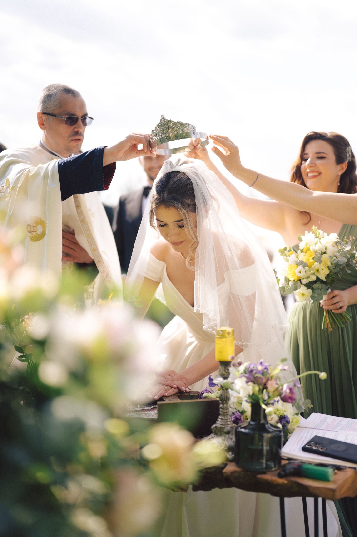 Guests help adjust a bride's veil during an outdoor wedding ceremony with floral arrangements and a bridesmaid in sage green.