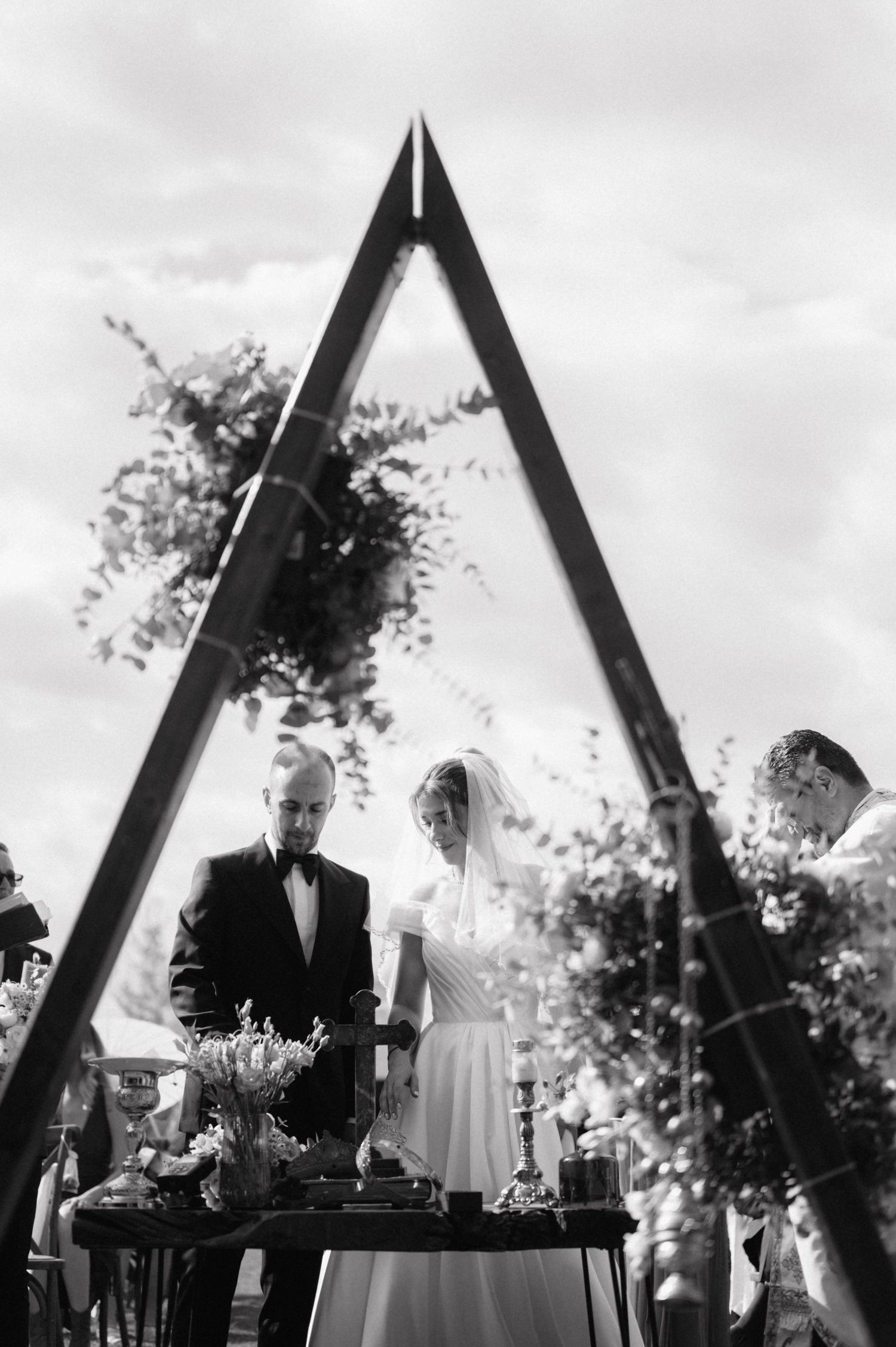 Black and white photo of a wedding ceremony beneath a triangular arch with floral decorations.