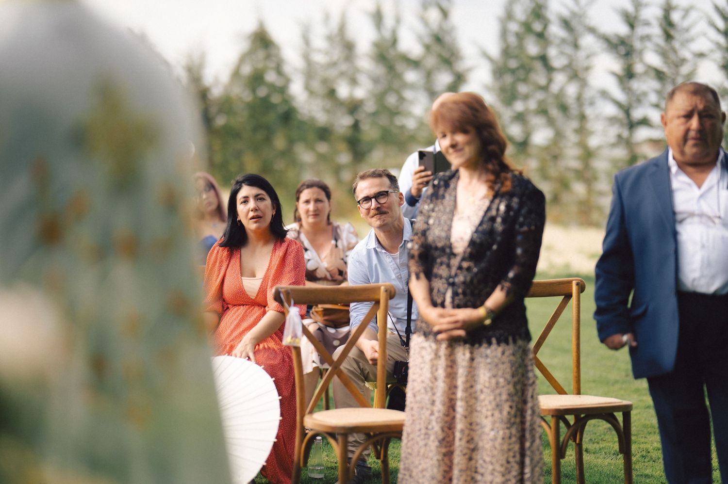 Wedding guests at an outdoor ceremony sitting and standing in wooden chairs with evergreen trees in background.