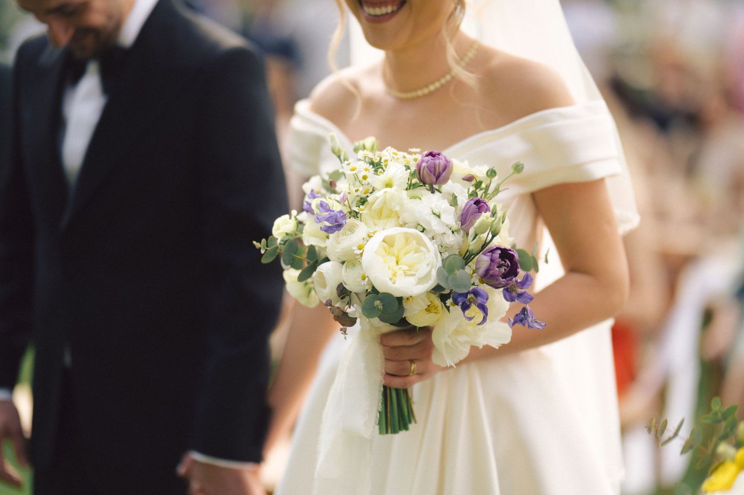 A close-up view of an ivory wedding bouquet with white roses, purple accents and greenery against a soft blurred background.