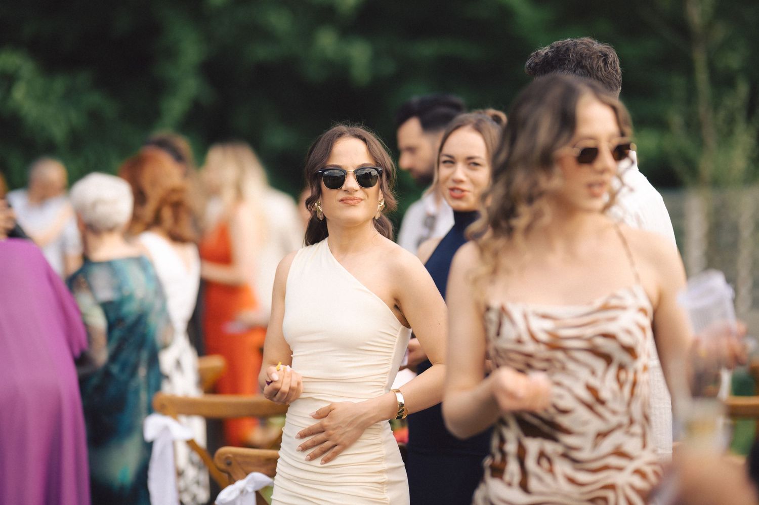 Wedding guests in formal attire gather outdoors, with people wearing sunglasses and summer dresses.
