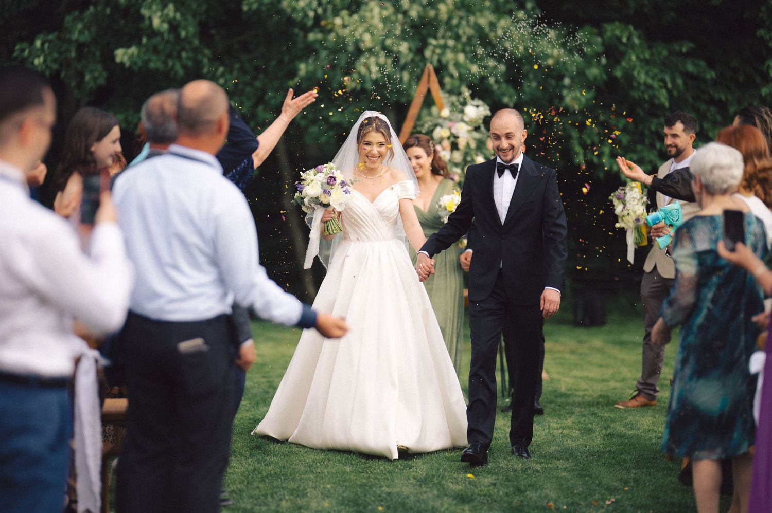 Newlywed couple celebrates while walking down outdoor wedding aisle as guests cheer with raised hands.