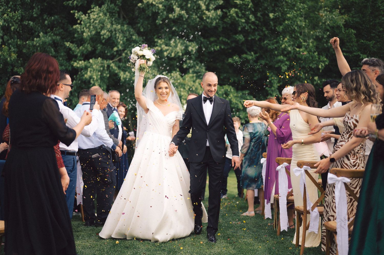 Newlyweds walk down outdoor aisle surrounded by cheering guests throwing confetti during summer garden ceremony.