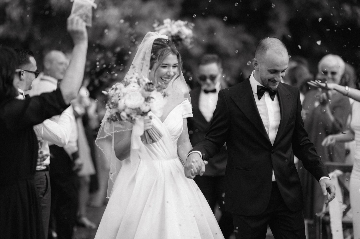 A joyful newlywed couple celebrates with guests as confetti falls around them at an outdoor wedding ceremony.
