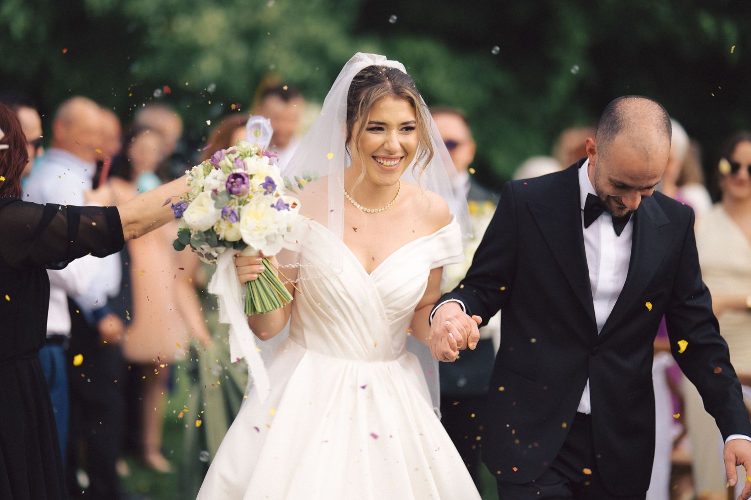 A joyful wedding celebration outdoors with confetti falling as the couple walks among guests.