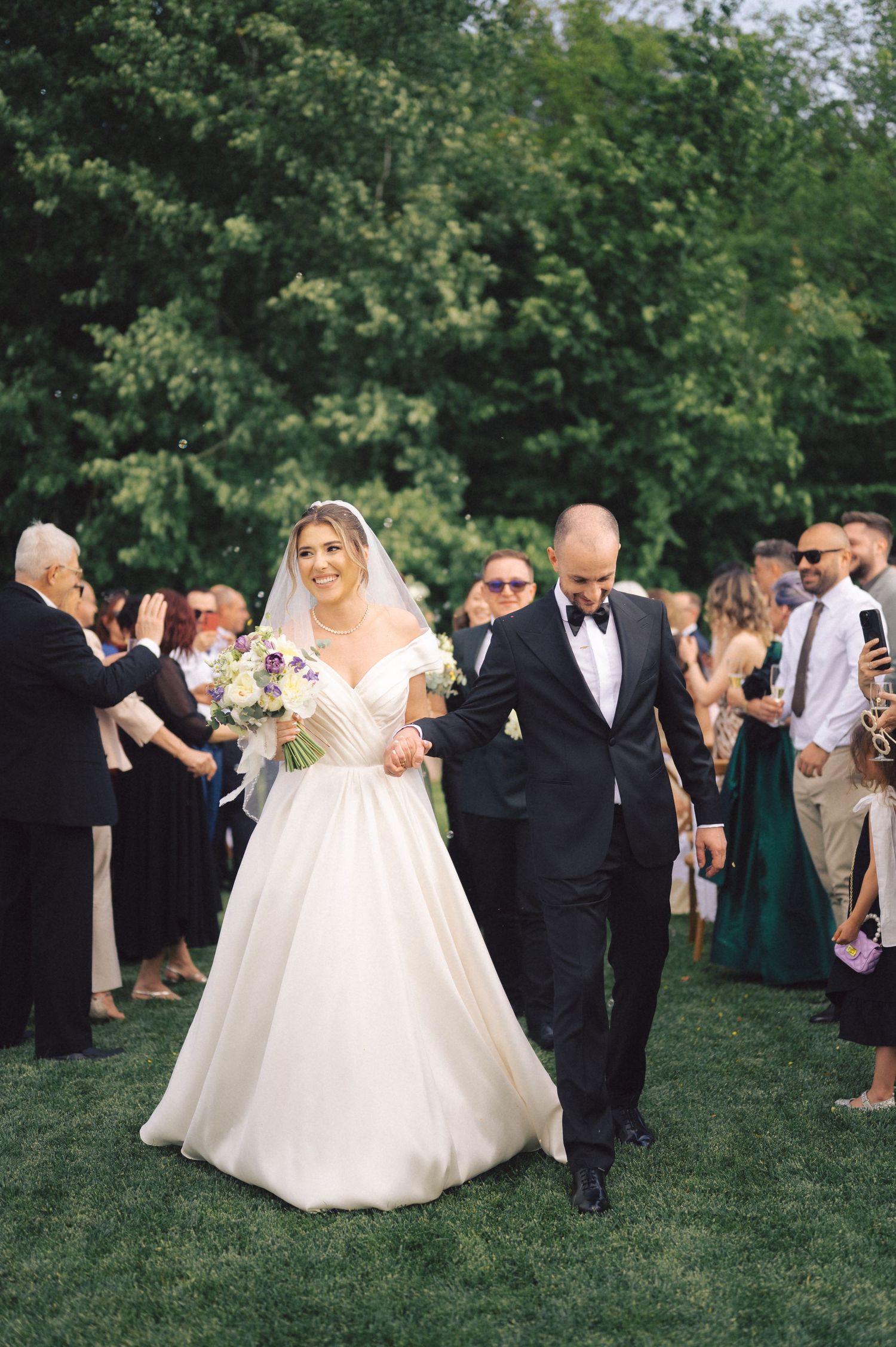 A couple walks down a grassy aisle at an outdoor wedding ceremony surrounded by guests and lush greenery.