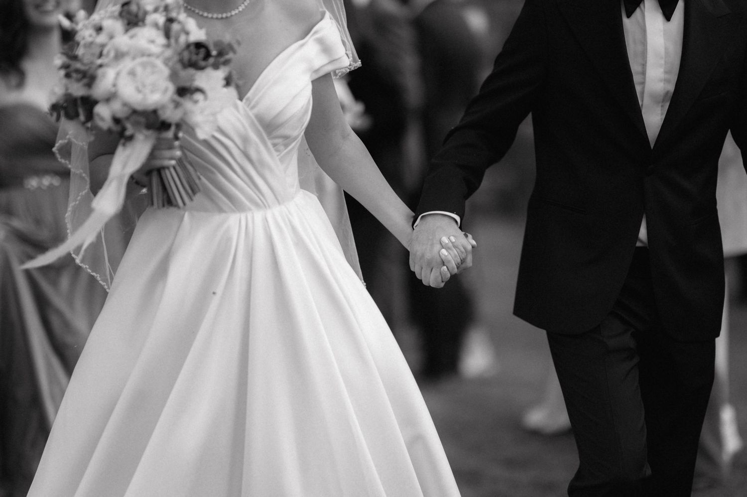 A black and white photo of a couple holding hands at a wedding, with the bride in white dress and groom in tuxedo.