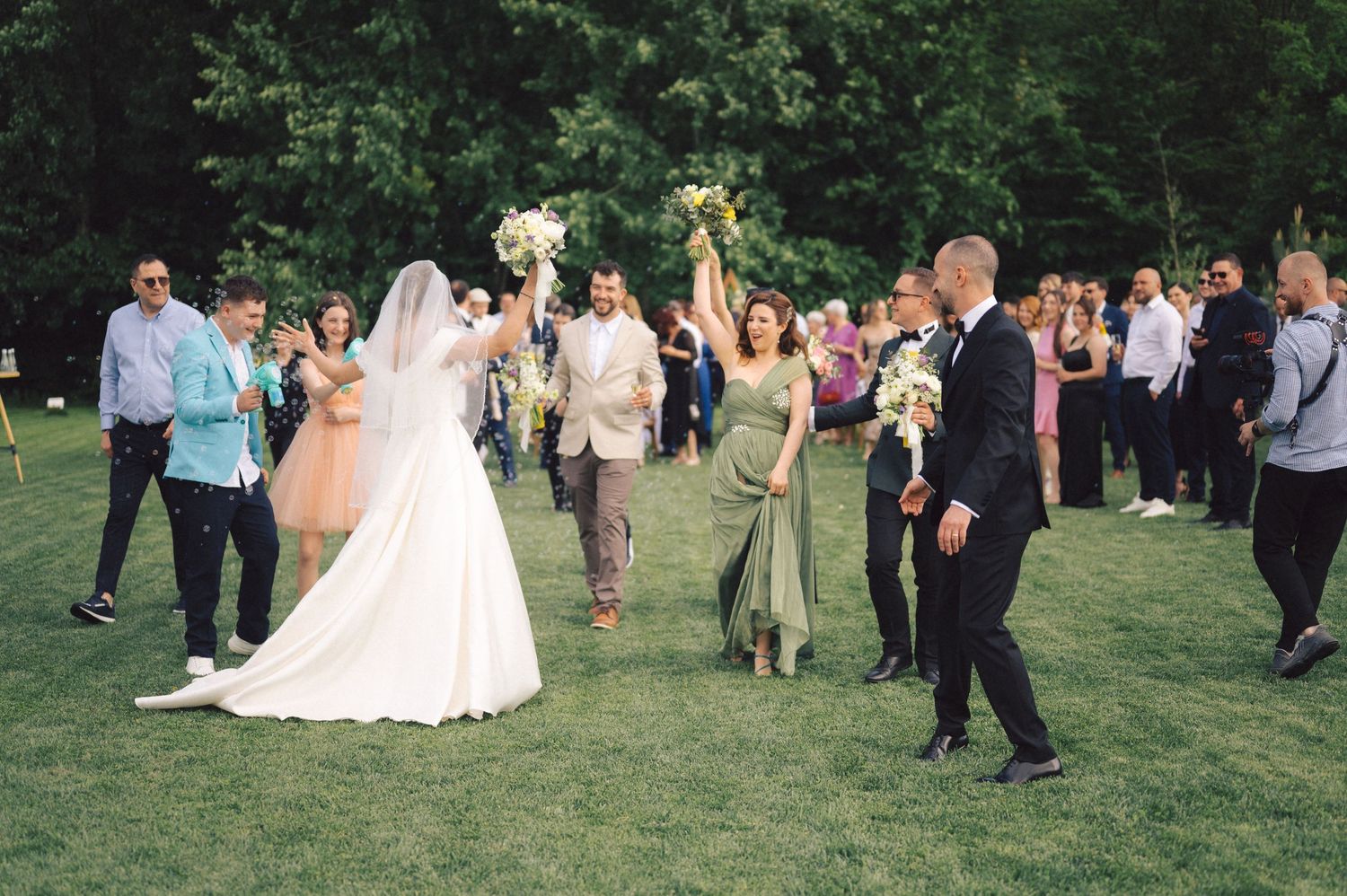 Wedding guests celebrate on a lush green lawn as someone in a mint green dress catches the bridal bouquet.