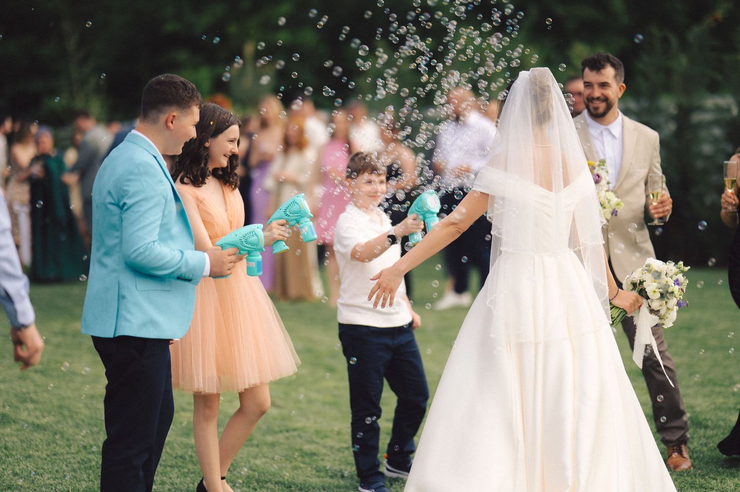 Guests shower newlyweds with bubbles during outdoor wedding celebration on grassy lawn.