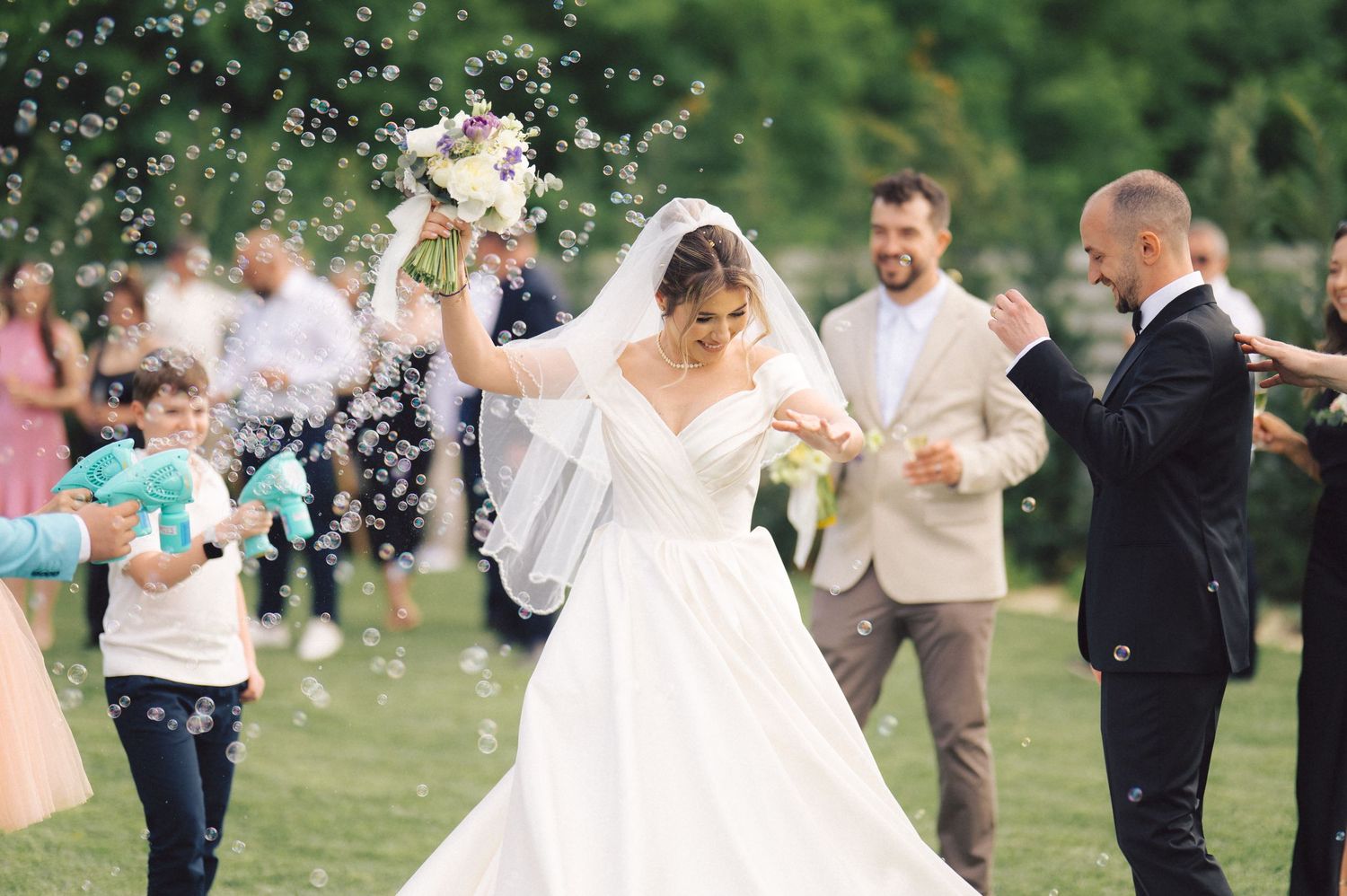 Wedding guests throw confetti as happy newlyweds celebrate on green lawn during outdoor summer ceremony.