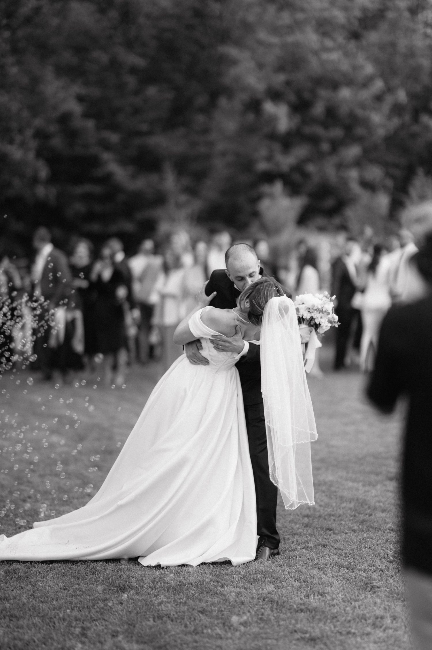 A couple shares a tender embrace during their outdoor wedding ceremony as guests look on in the background.