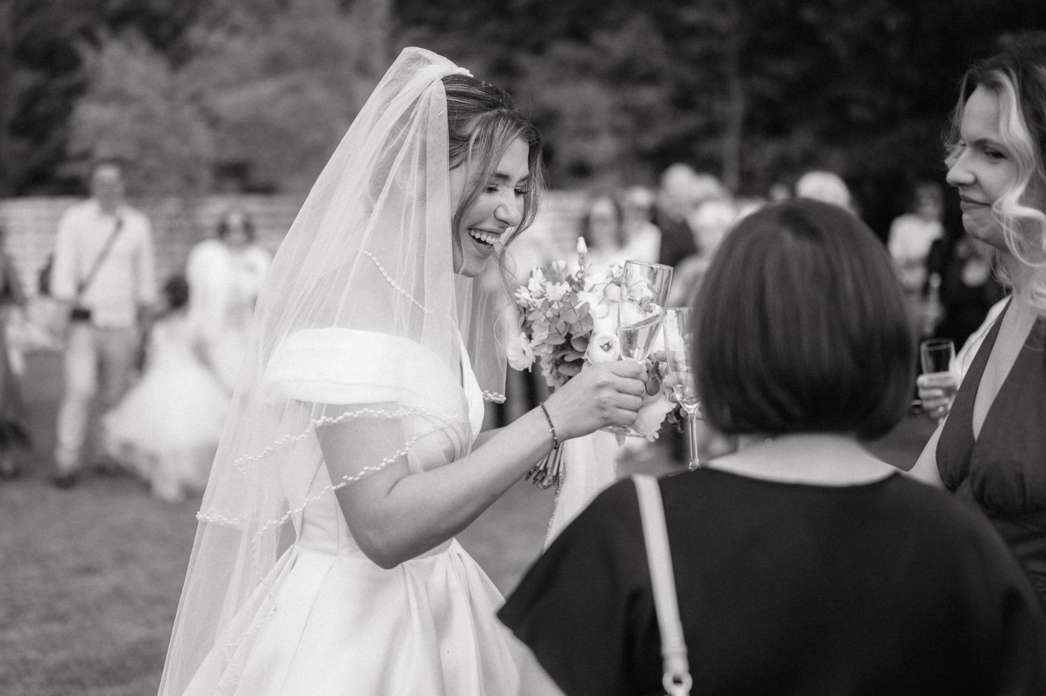A black and white candid photo shows a bride in a white gown and veil laughing during an outdoor reception.