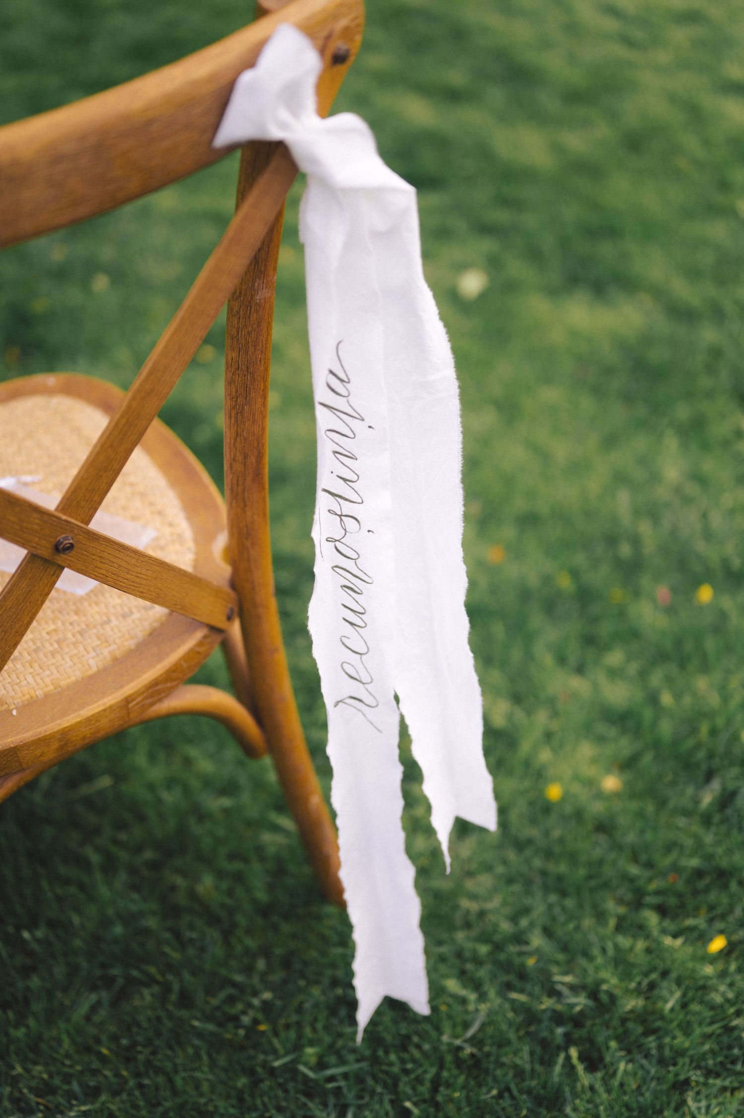 White ribbon tied to a wooden bistro chair on grass with decorative script writing.
