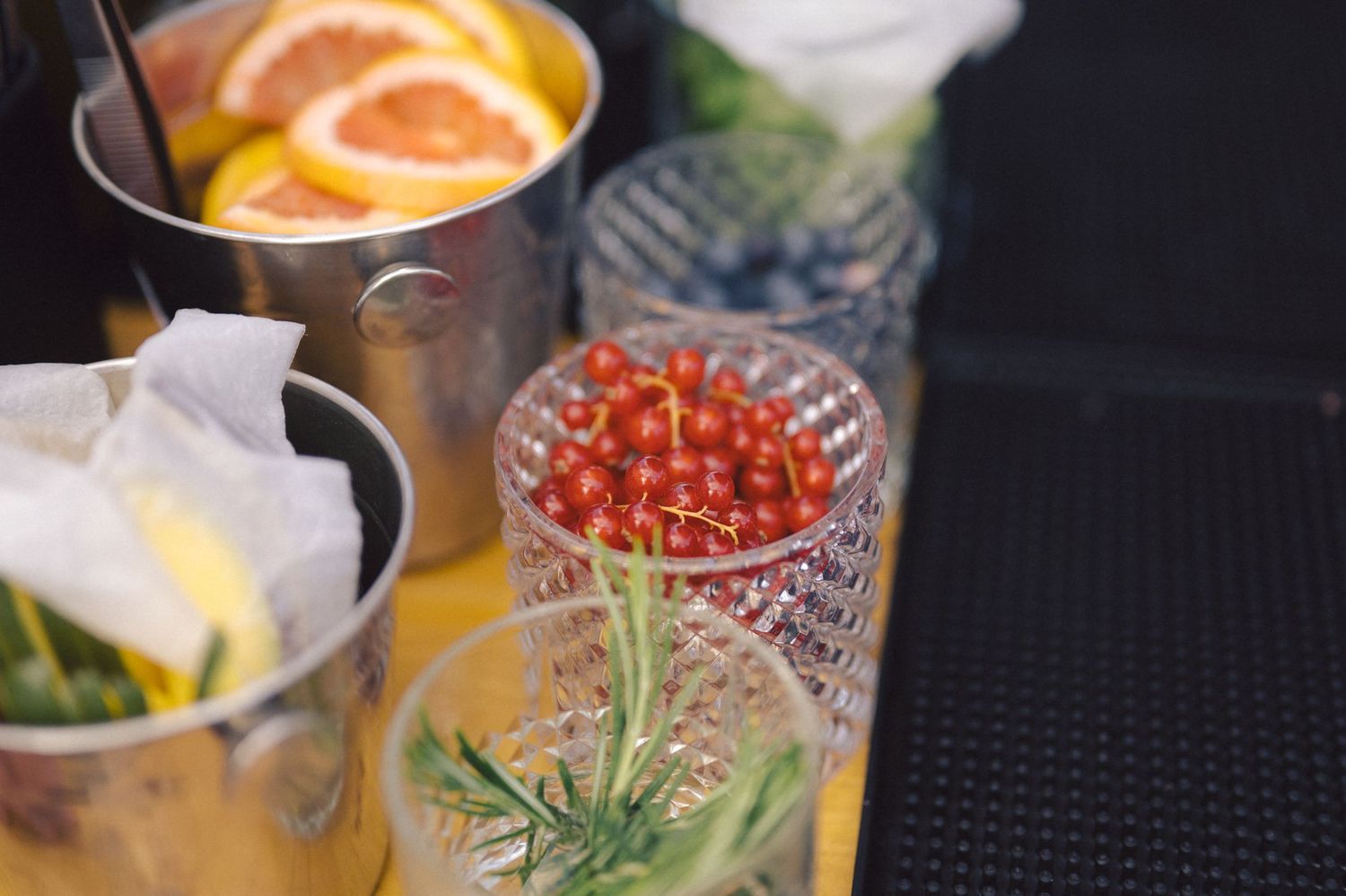 Close-up of cocktail glasses with citrus garnishes and red berries on a dark bar surface.