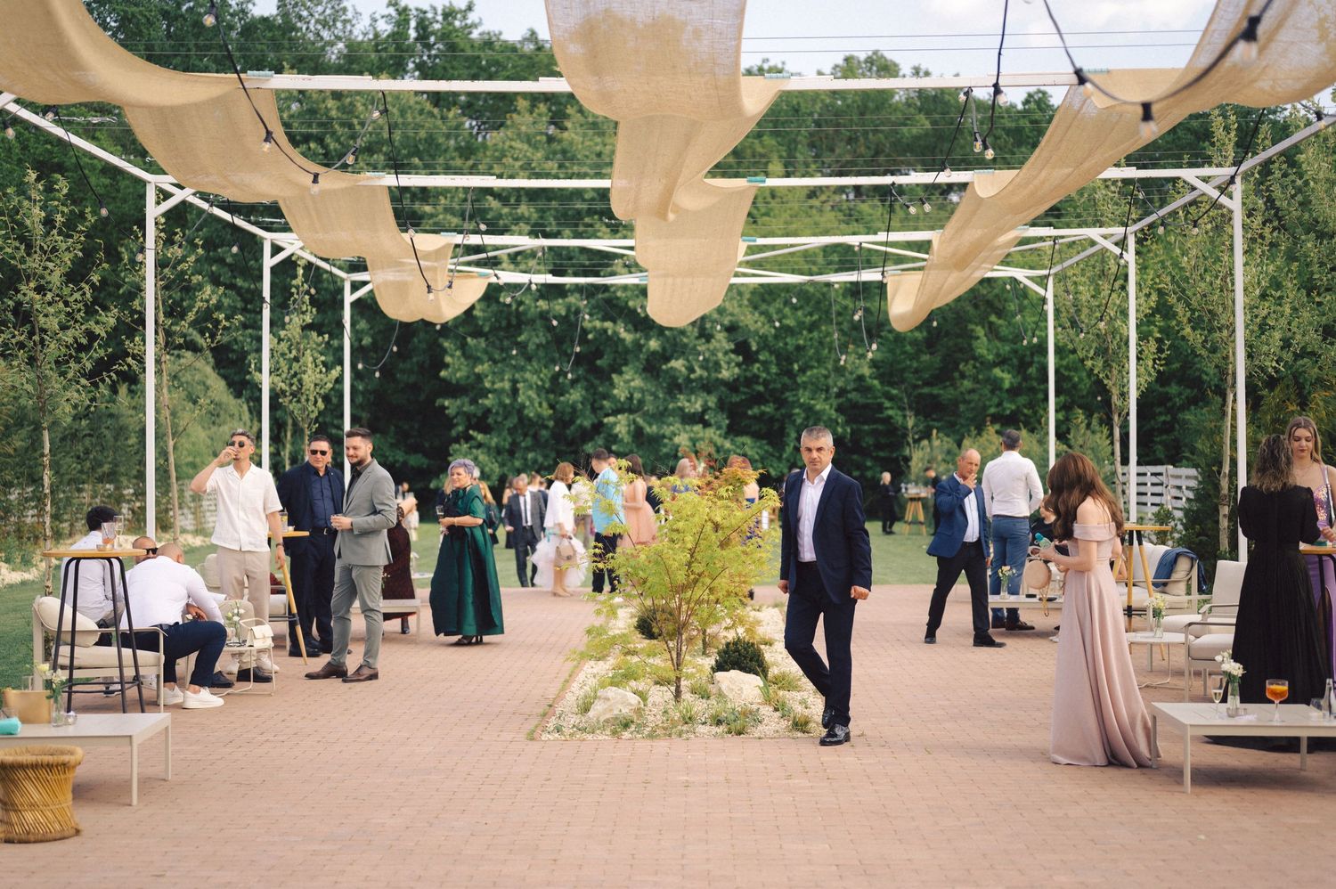 Outdoor wedding reception with elegant canopy and guests mingling in a garden setting under wooden beams.