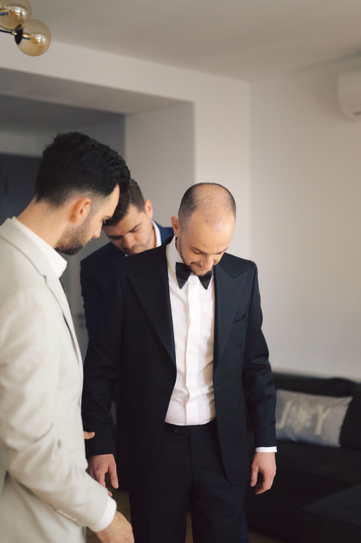 Three men in formal attire huddle together while looking down in a minimalist interior setting.