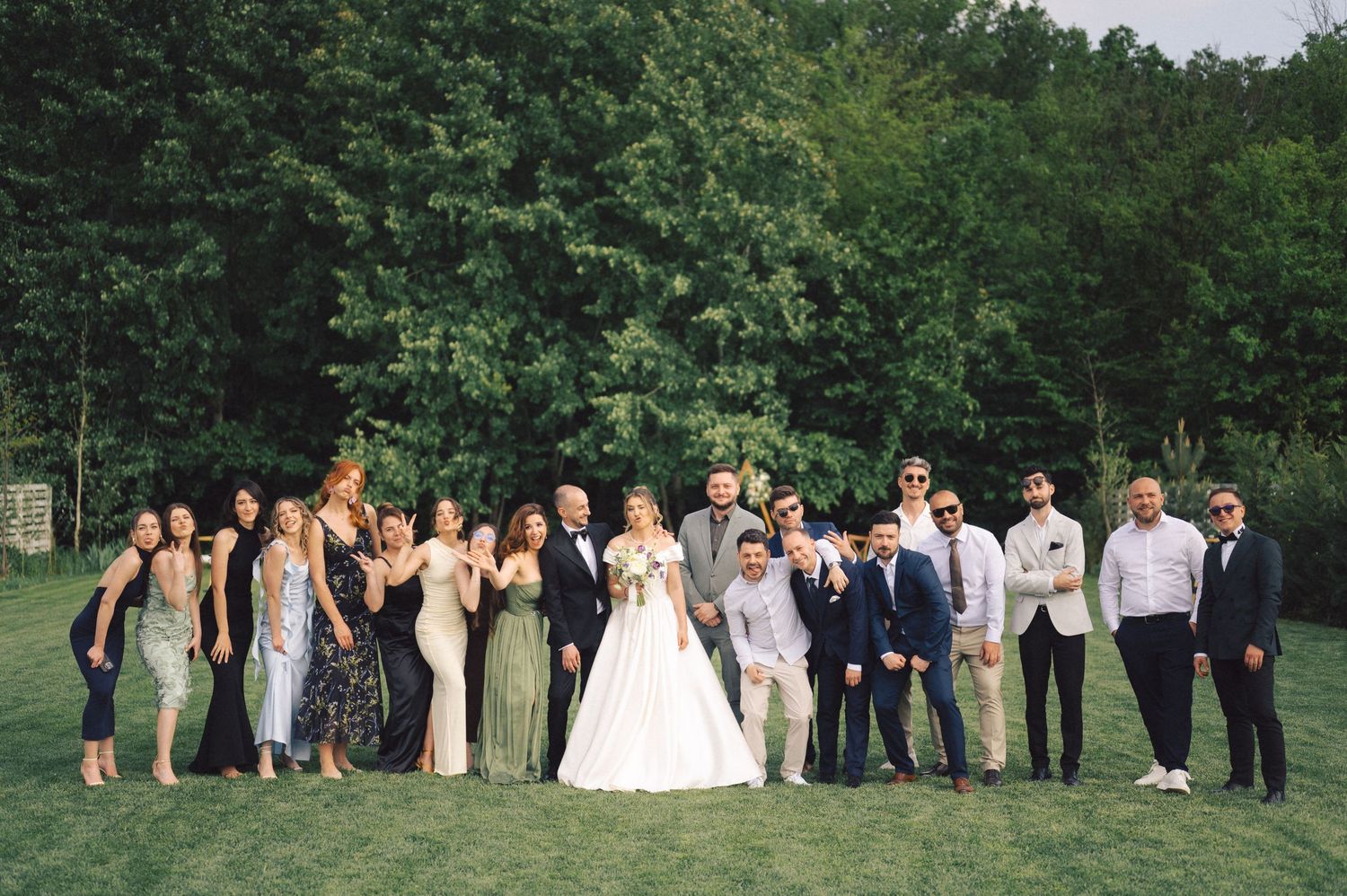 Wedding party poses together on a lush green lawn surrounded by trees during an outdoor summer celebration.