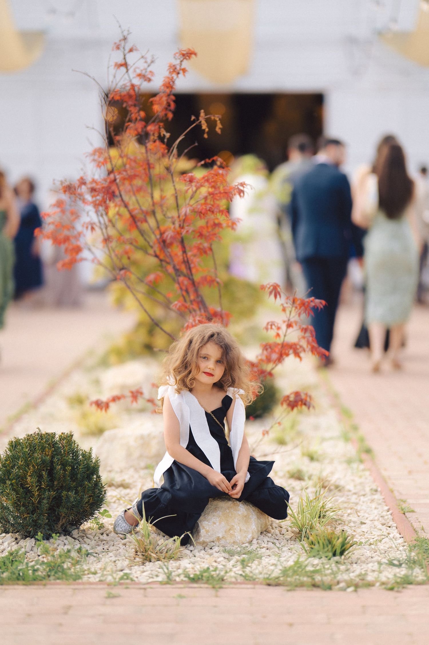 A young child in a black and white dress sits beneath a blooming red maple tree while others gather in the background.