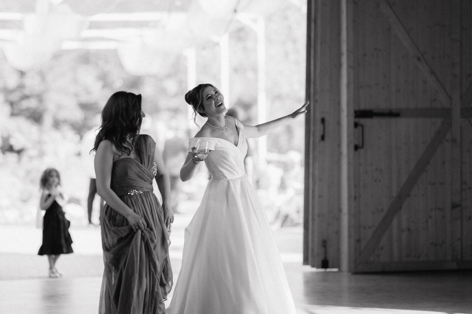 Two women in elegant dresses share a joyful moment at a wedding venue while a small figure appears in background.