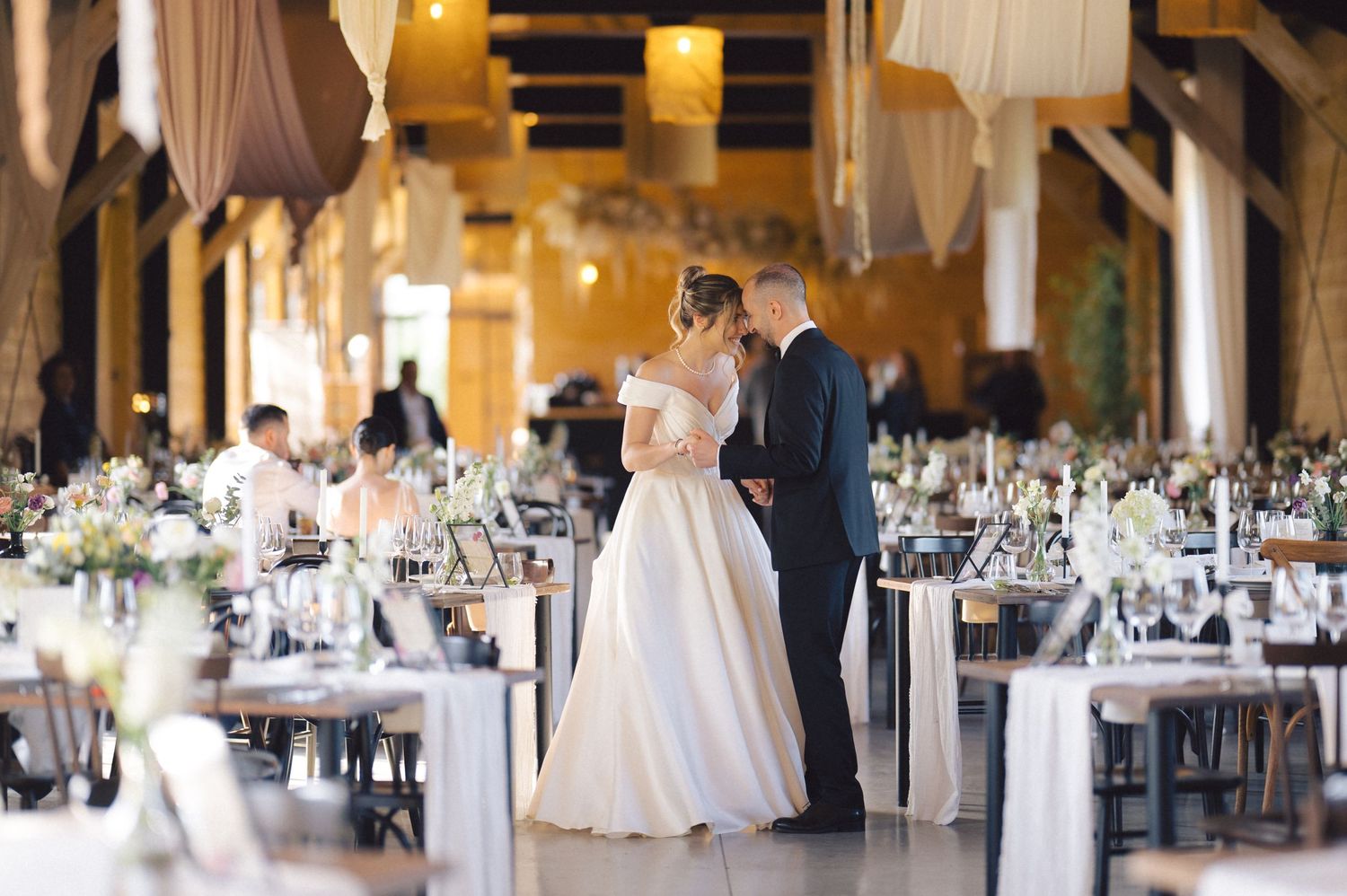 A romantic wedding reception setup in an elegant rustic barn venue with tables adorned in white linens and floral centerpieces.
