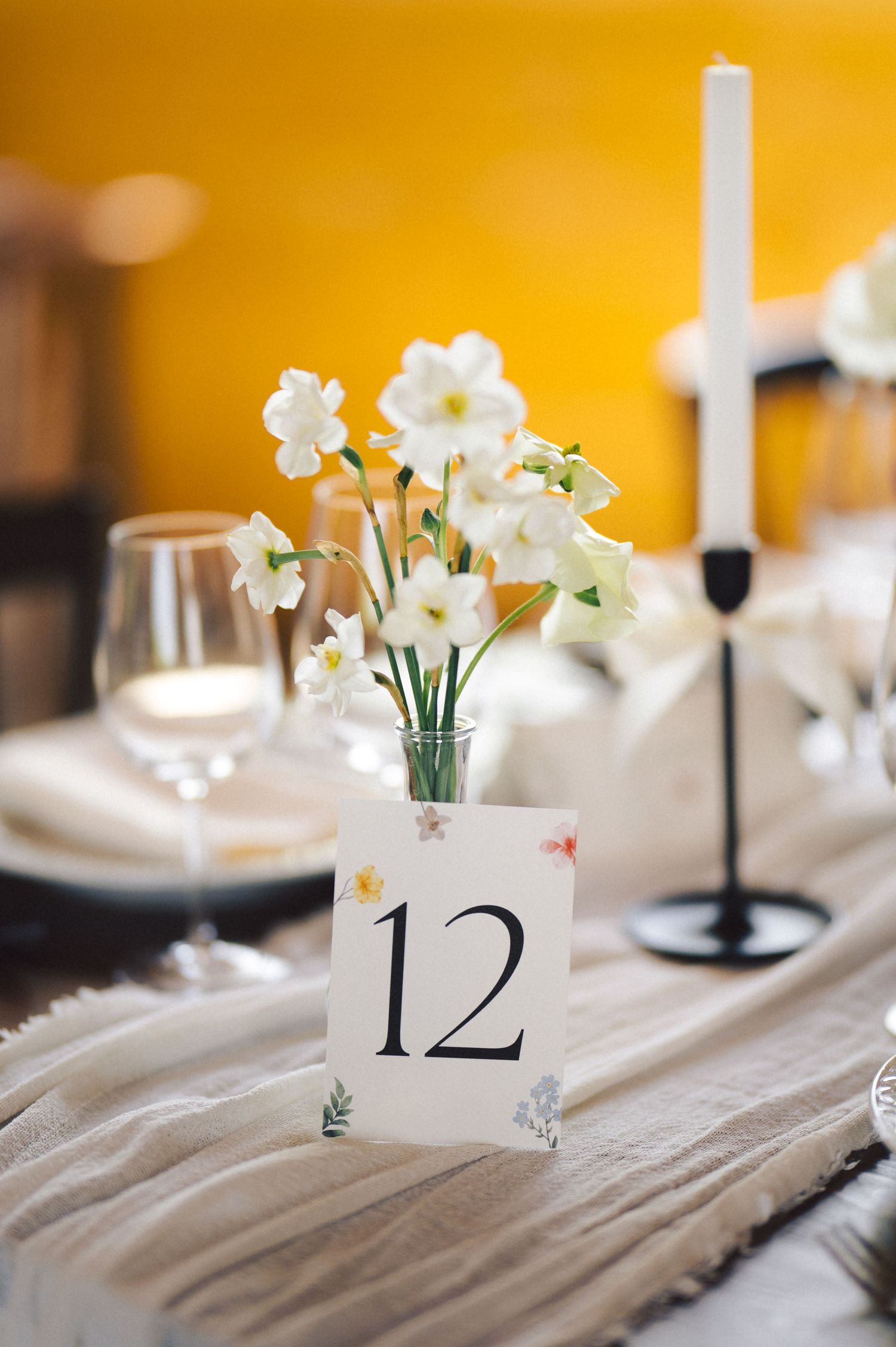 Elegant table setting with white narcissus flowers in vase, table number 12 card, and candlestick on neutral tablecloth.