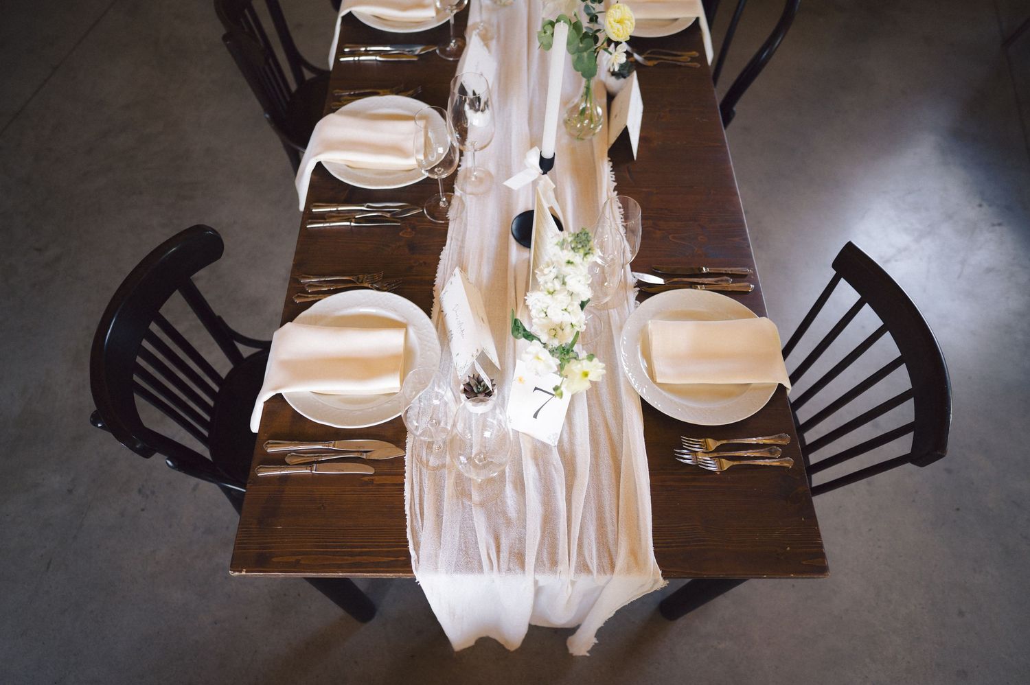 Elegant table setting with white runner and place settings on dark wood table with black dining chairs.