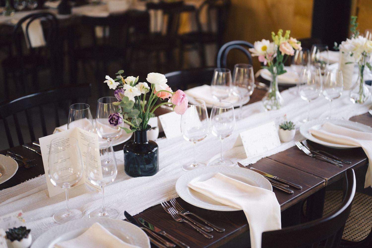 An elegant table setting with white linens, dark wood tables, and small floral centerpieces in black vases.