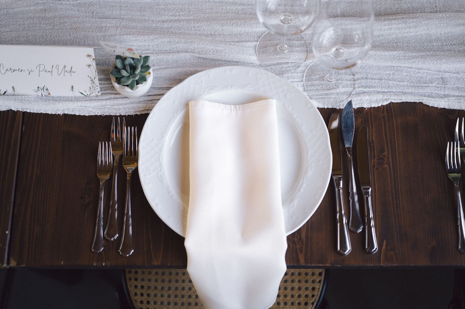Formal place setting with white plate, napkin and multiple utensils on dark brown table runner.