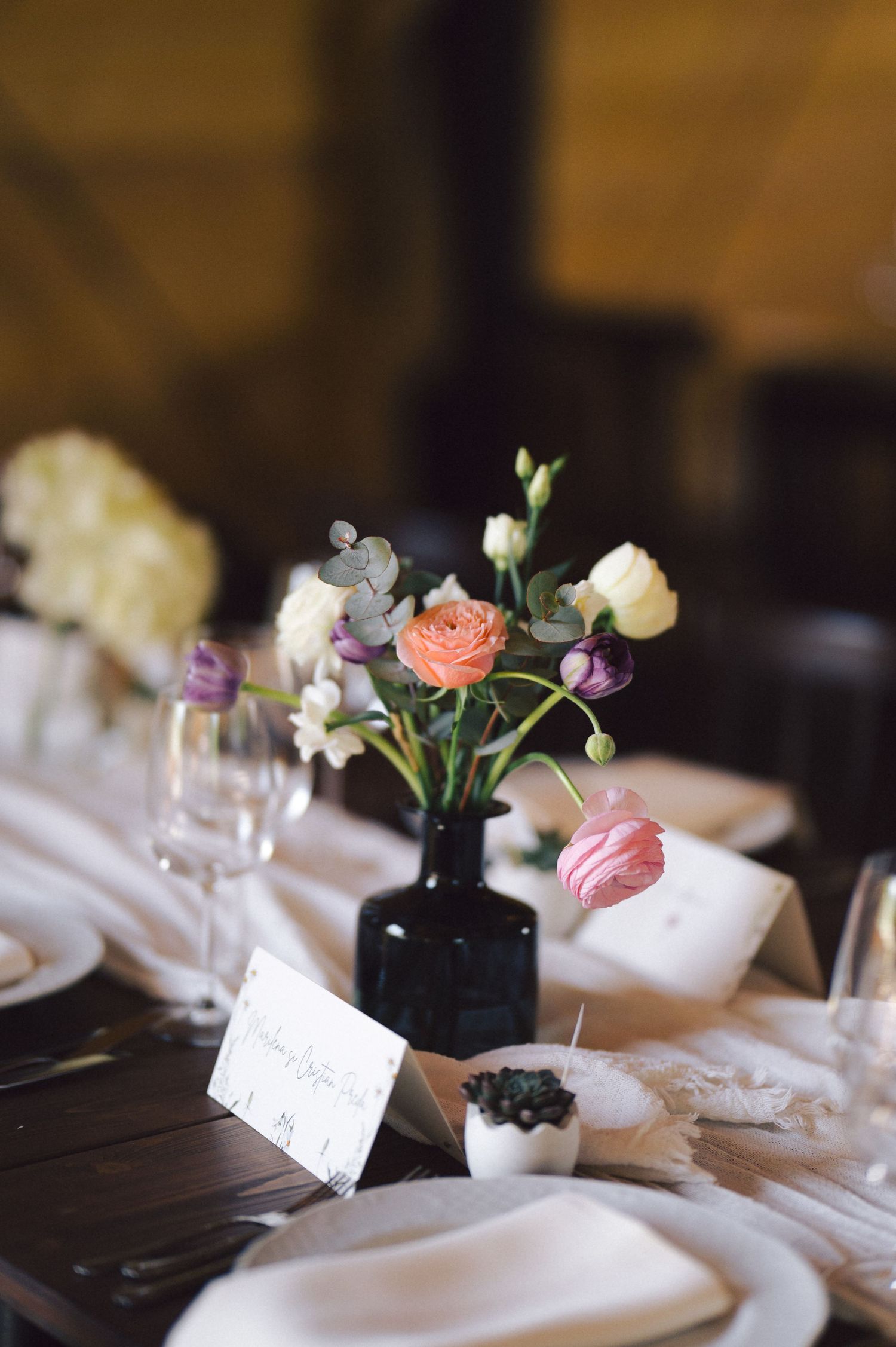 A small bouquet of colorful flowers in a black vase adorns a table setting with white linens and elegant place settings.
