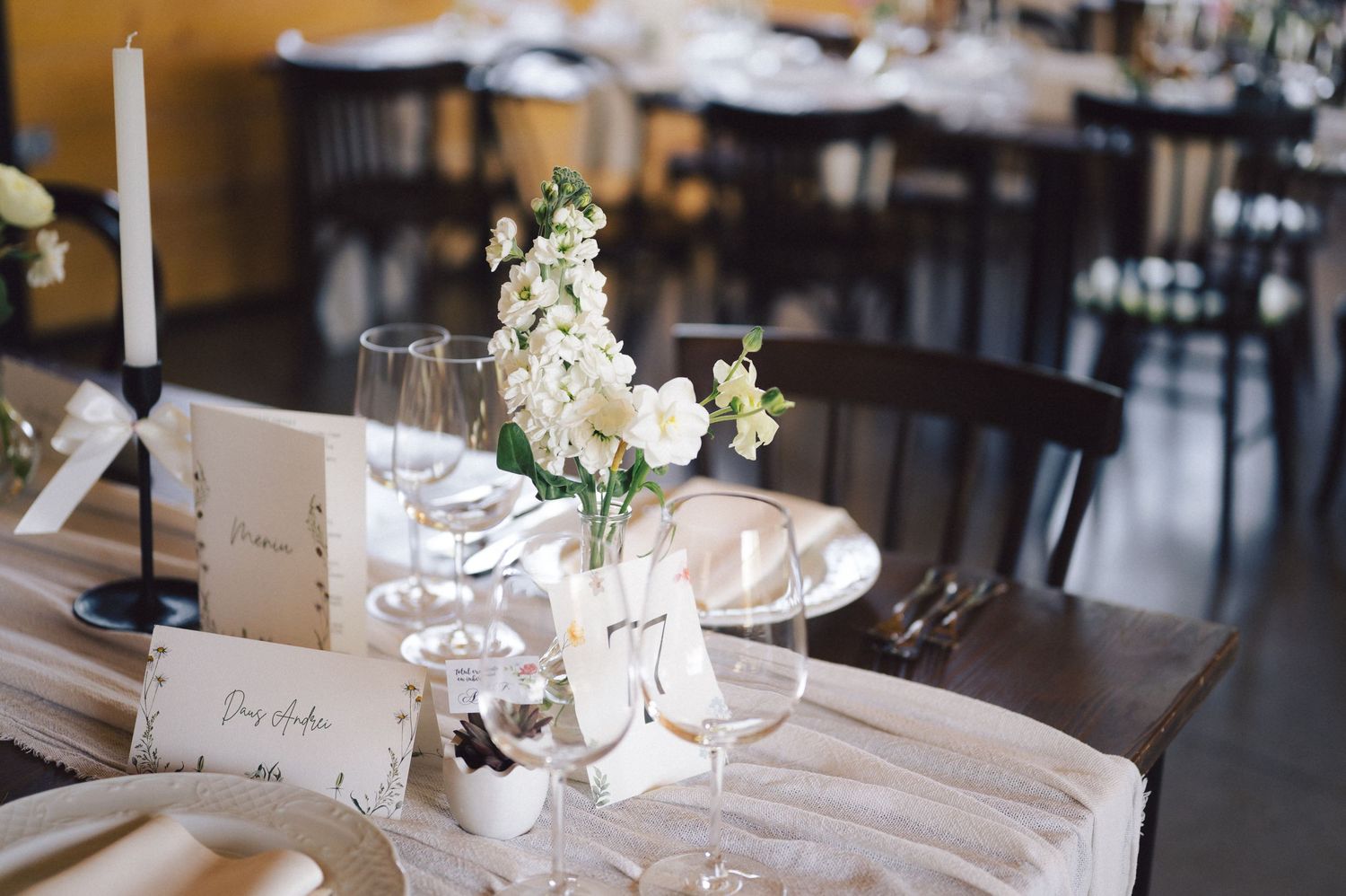 An elegant wedding table setting with white floral centerpiece, wine glasses and place cards on a light-colored tablecloth.