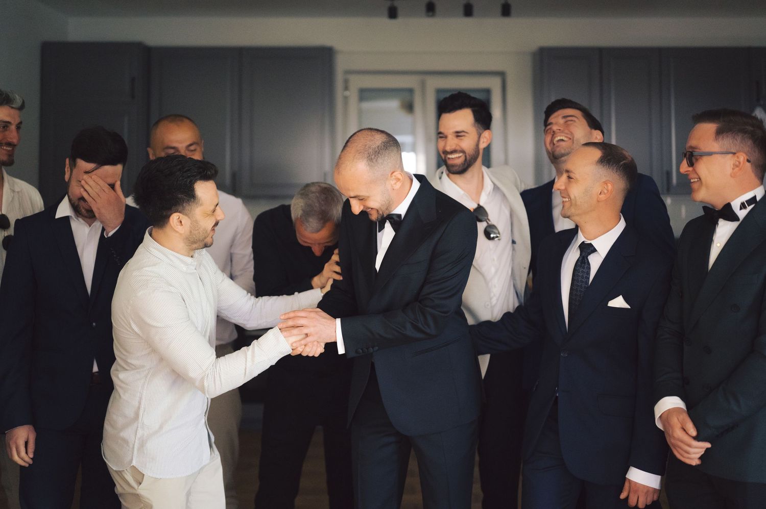 A groom in a white suit and guest in black tuxedo shake hands during an elegant indoor wedding celebration.
