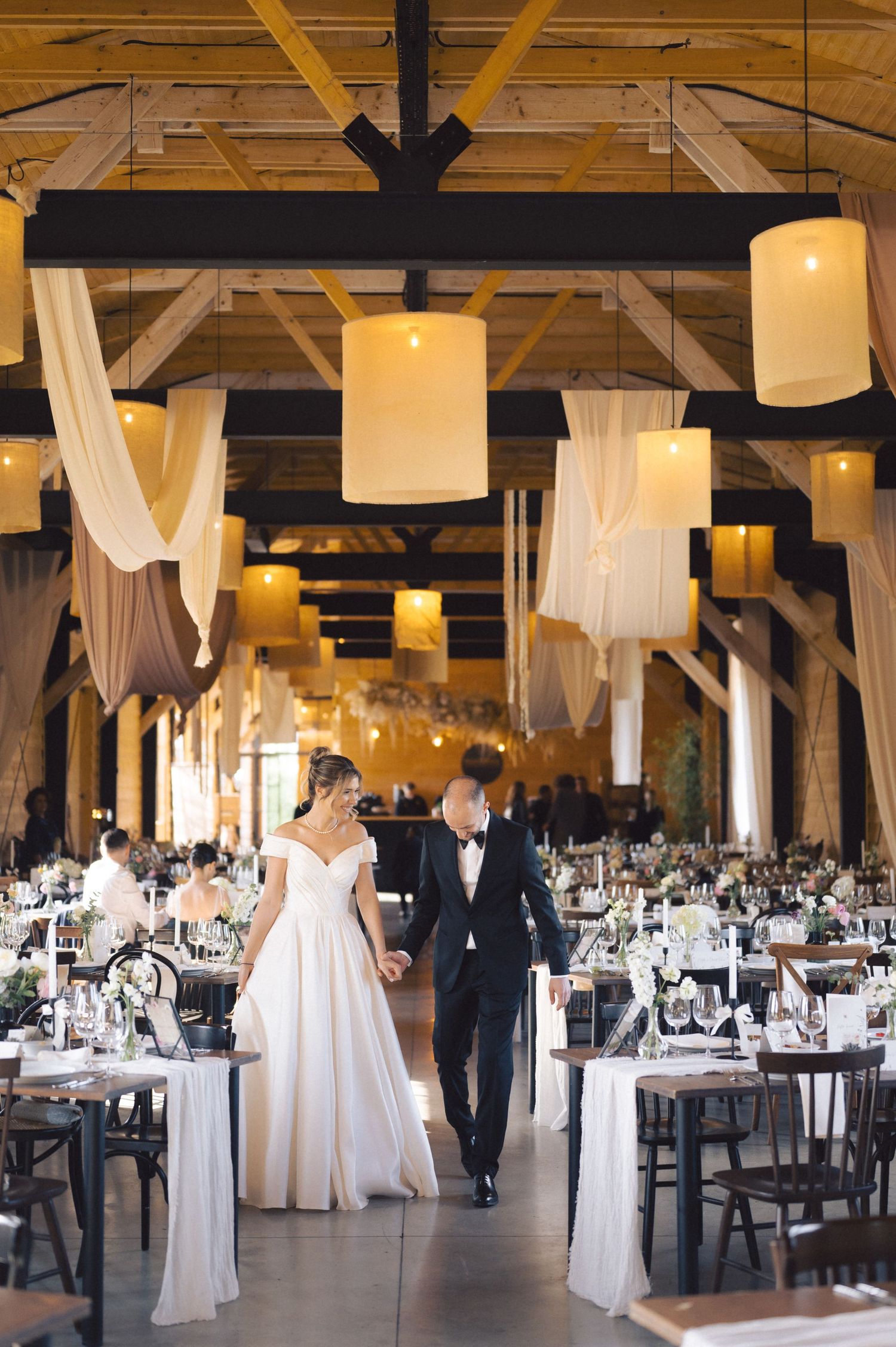 Elegant wedding reception venue with rustic wooden beams, hanging fabric drapes and glowing cylindrical lantern lighting.