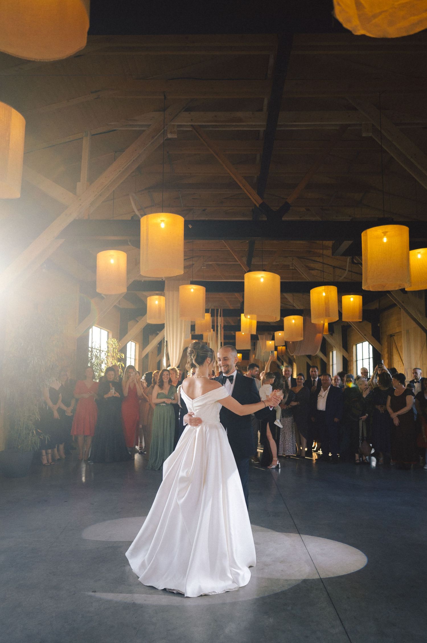 A couple dancing under glowing lanterns in a rustic venue with guests watching in the background.