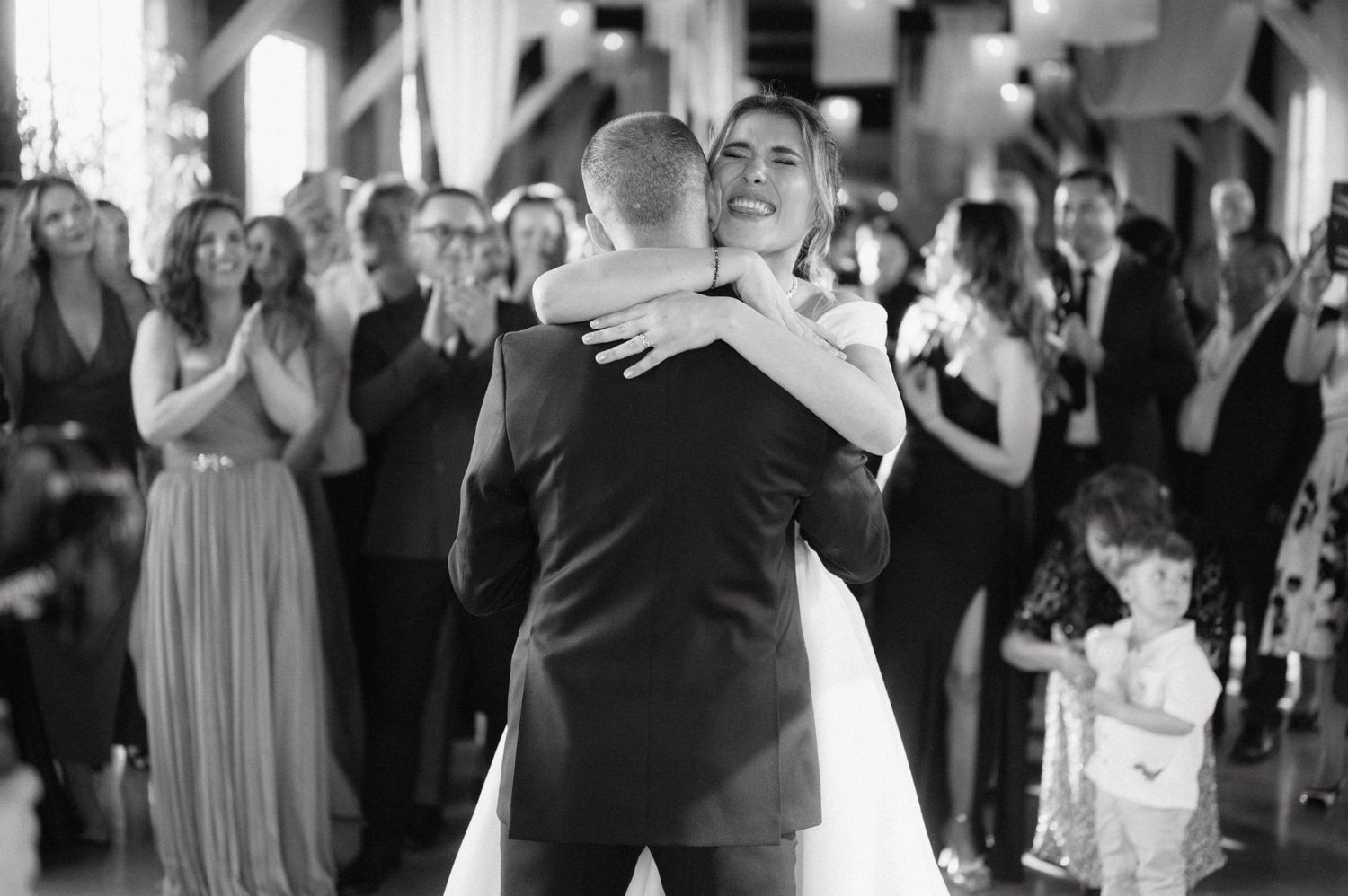 A couple shares a heartfelt embrace during their first dance at a wedding reception while guests watch and applaud.