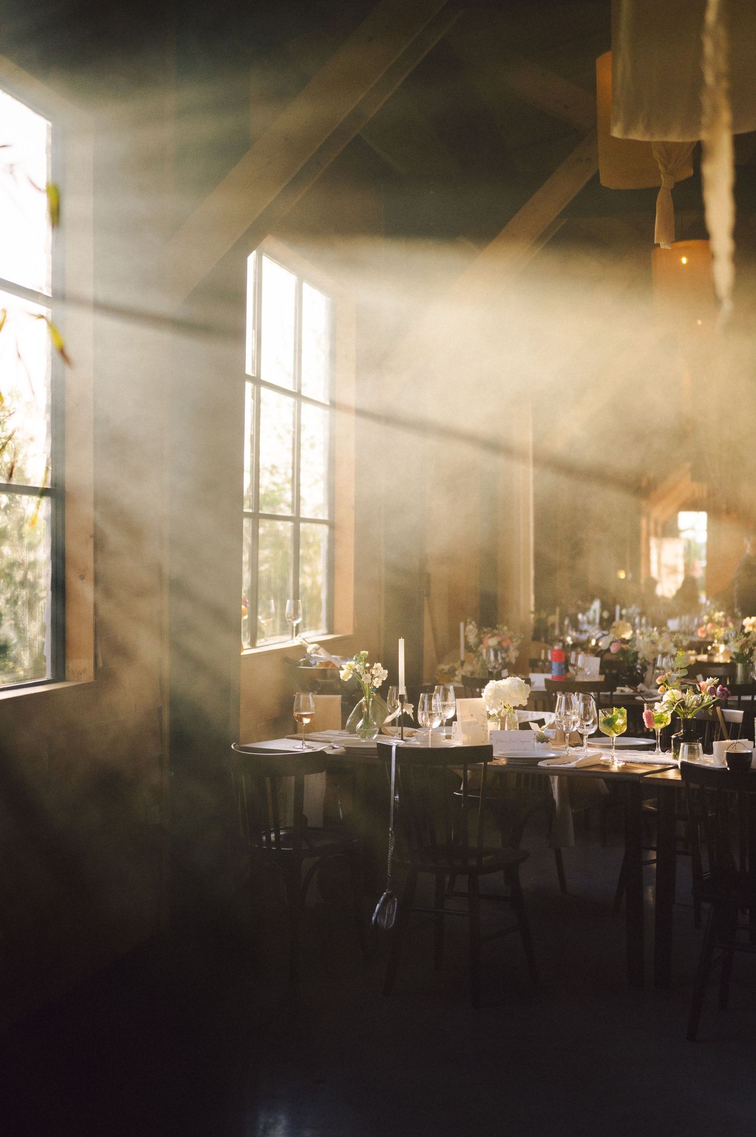 Sunlight streams through windows illuminating an elegant rustic dining room setup with tables and chairs.