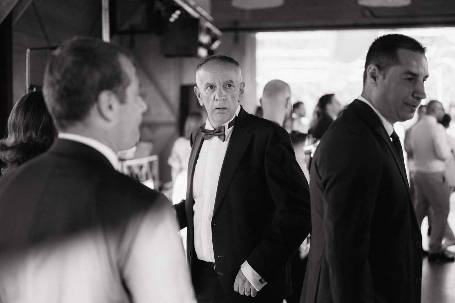 Men dressed in formal attire stand together at an elegant social gathering in black and white photography.