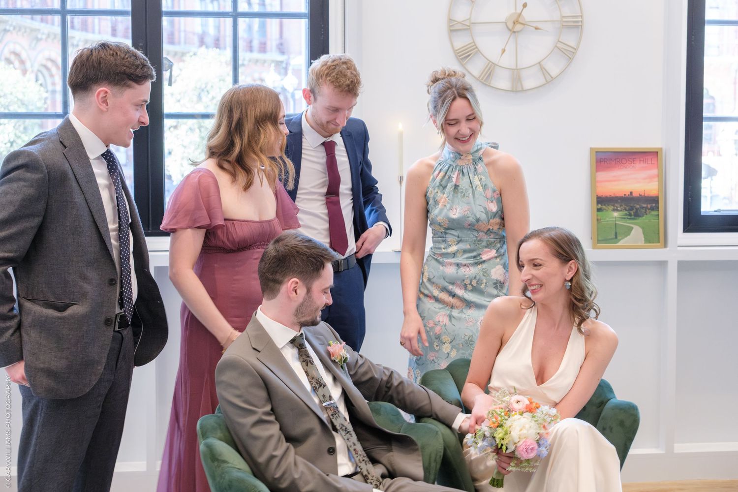 Wedding party gathered inside bright room with large windows and decorative clock on wall during celebration.