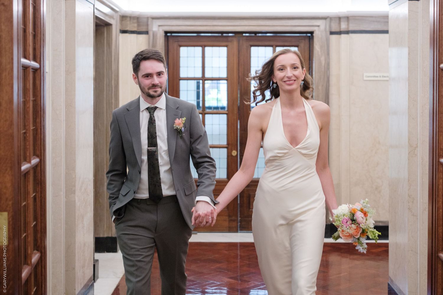 A newlywed couple joyfully walks down a hallway holding hands while wearing a gray suit and white wedding dress.