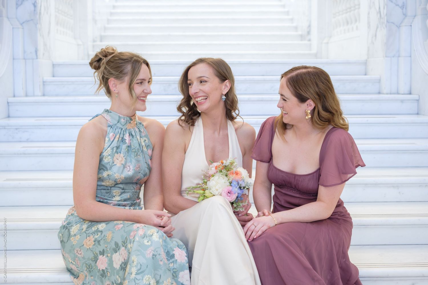 Three women in formal dresses share a joyful moment while sitting together on white marble steps.