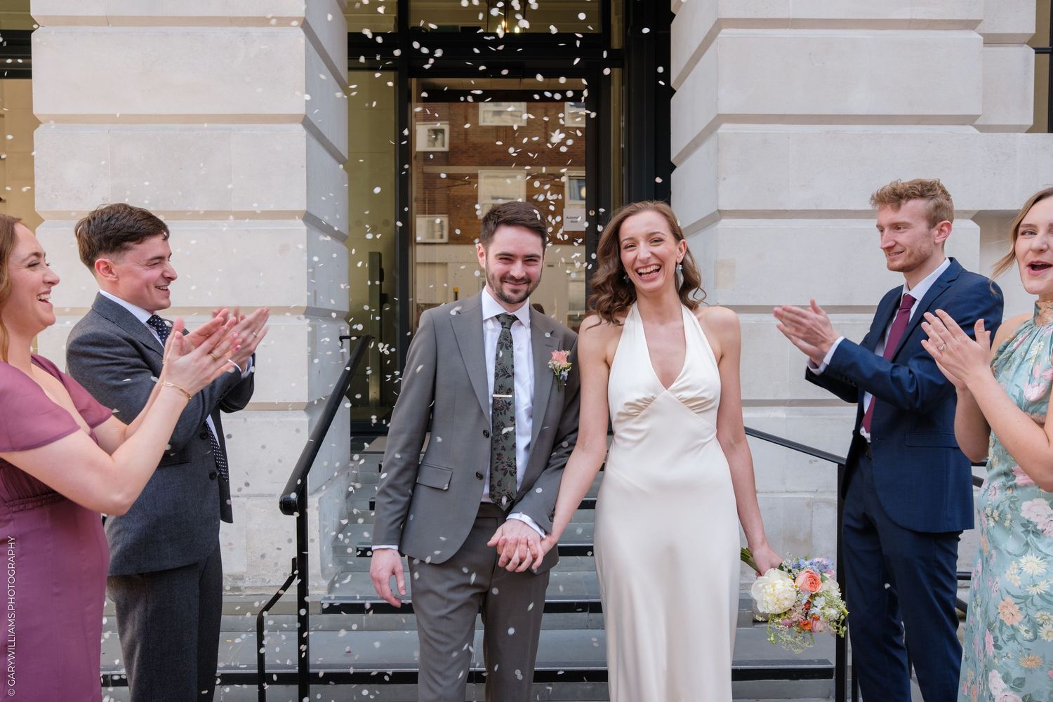 Wedding guests clap and celebrate as a couple in elegant wedding attire exits a building with festive decorations.