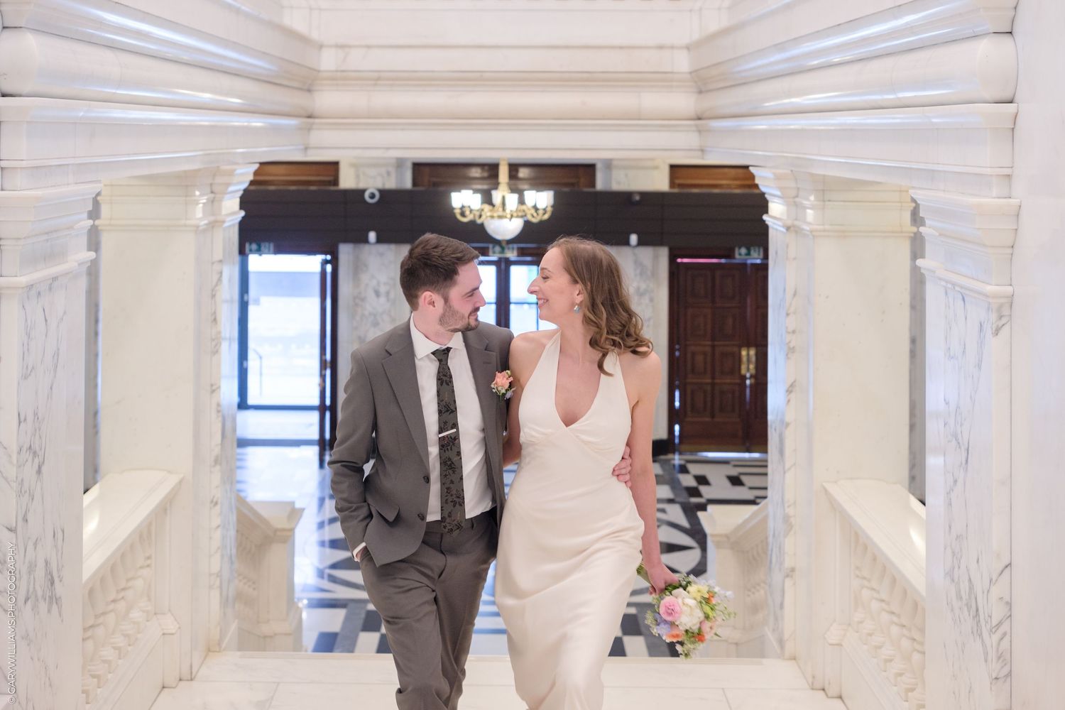 A couple in wedding attire shares an intimate moment while sitting on white marble steps in an elegant venue interior.