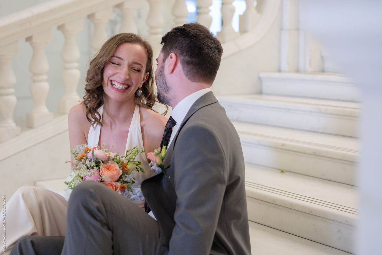 Happy couple sits together on elegant white marble staircase with bride holding colorful wedding bouquet.