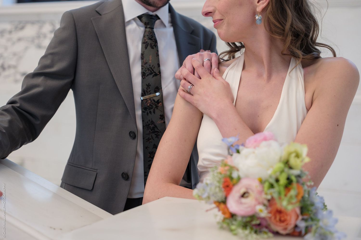Close-up view of a wedding bouquet with pastel flowers held near a gray suit and white dress.