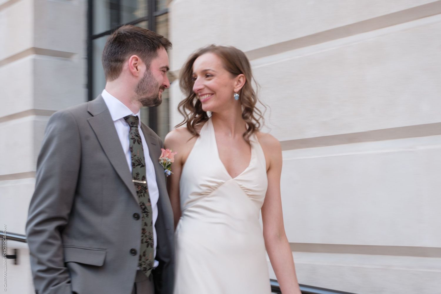 A bride in a white halter dress laughs with someone wearing a grey suit outside a building with stone walls.