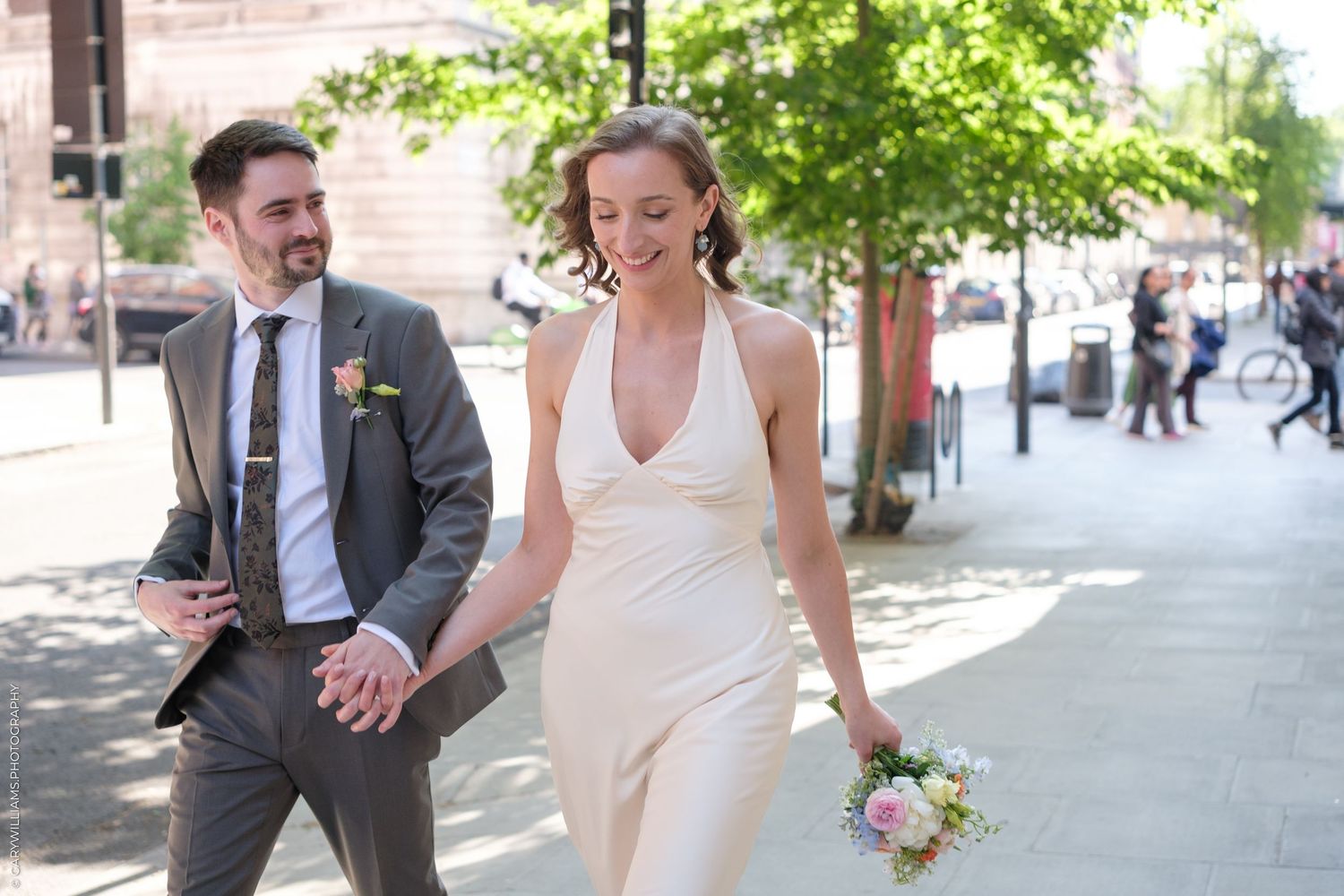 A joyful couple laughs while walking down a sunny city street, the bride in a white halter dress holding a small bouquet.