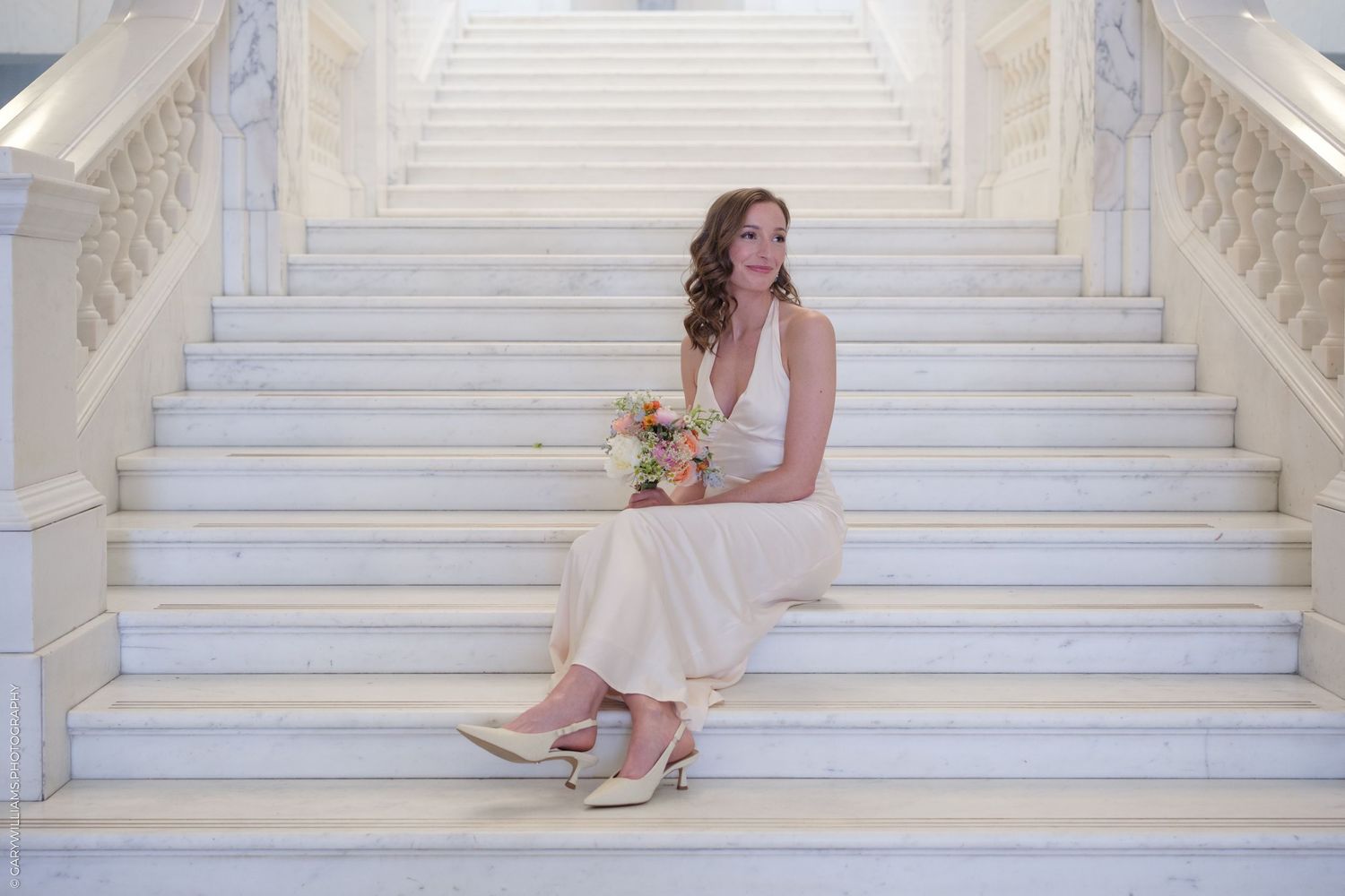 A person in a flowing blush gown sits gracefully on elegant white marble stairs while holding a coral bouquet.